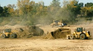 Three yellow tractors with loaders moving sand on an empty lot with green trees in the background.