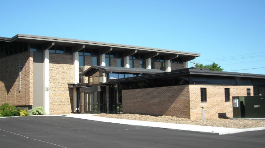 Bi-level brown brick building with glass entrance on lower half and asphalt parking lot in front.