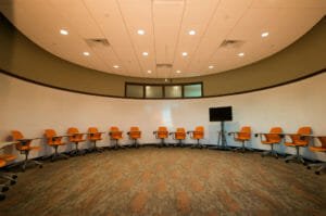 Round room with green and orange carpet tiles, orange desk chairs on back wall with round whiteboard on entirety of rounded wall.