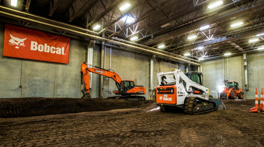Large interior space with dirt floor and concrete walls and overhead metal roof and exposed beams with Bobcat equipment and Bobcat banner on back wall.