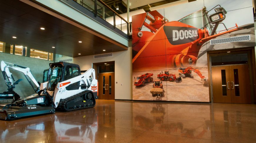 Concrete speckled flooring with bobcat machinery to back left and floor-to-ceiling paneled accent wall with picture of Doosan equipment on back wall with second-floor mezzanine overlooking.