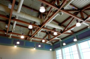 White metal ceiling with brown beams and exposed HVAC systems with hanging pendant lights throughout.