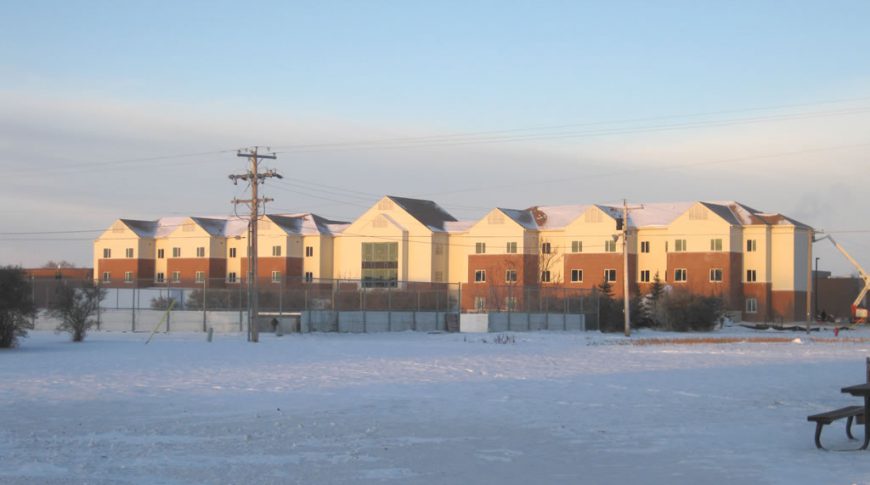 Far away view of entire F08 dormitory building on the Minot Air Force Base on a winter day. Snow in foreground with cream and red-bricked three-story dormitory building in background.