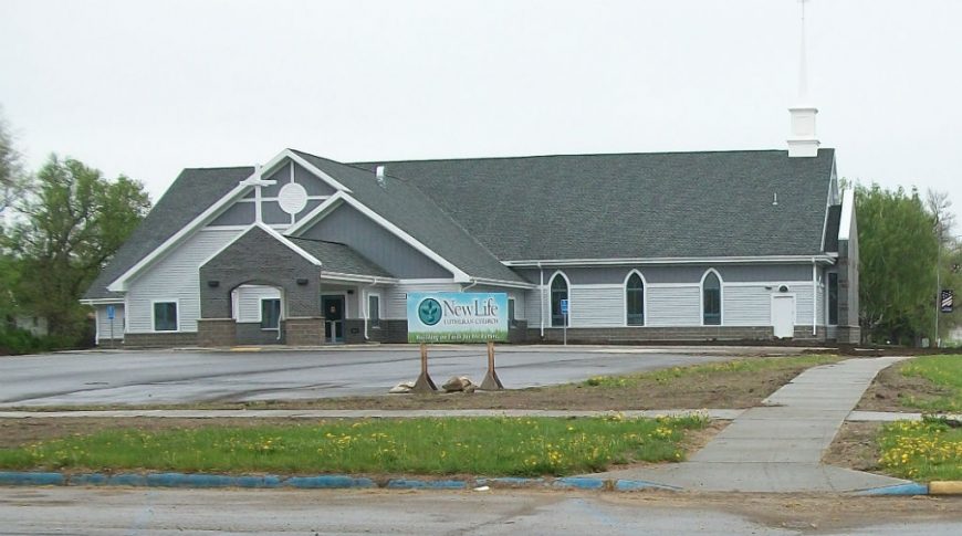 Road view of light and dark gray church with white steeple cross and cross over carport.