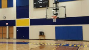 Half-court view of basketball hoop with blue padding behind and blue and yellow painted accents on walls with the words "Rothsay Tigers" painted in gold on the floor.