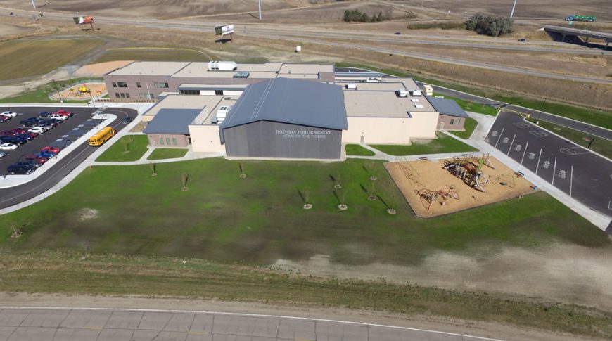 Side aerial view of Rothsay Public School with playground area to right foreground, school to center and parking lot to the left. Highway can be seen in background.