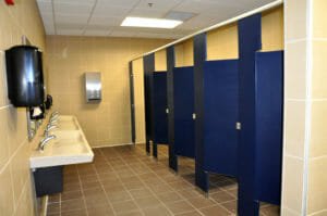 Public school restroom with bright blue stalls to the right side, beige tiles throughout the floor and walls, and sink stations to the left side.