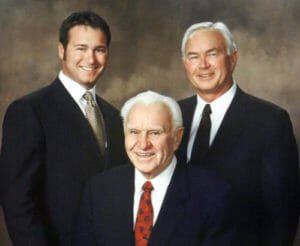 Three generations of Comstock men with black suites, white-collared shirts and ties on brown backdrop
