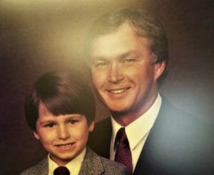 Father and young son sitting for portrait with suits and ties on brown backdrop