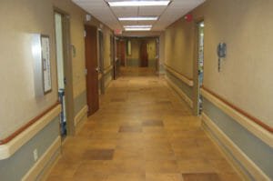 Hospital hallway with beige tiled floor underneath and blue and beige colored walls with chair ledge on each side and doorways to rooms on each side.
