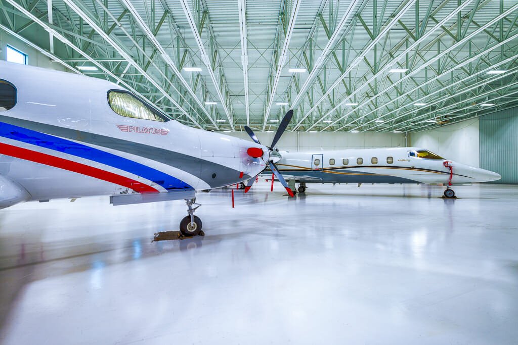 Interior of hangar building with light white floors and white metal ceiling overhead with fluorescent lights and private jets throughout.