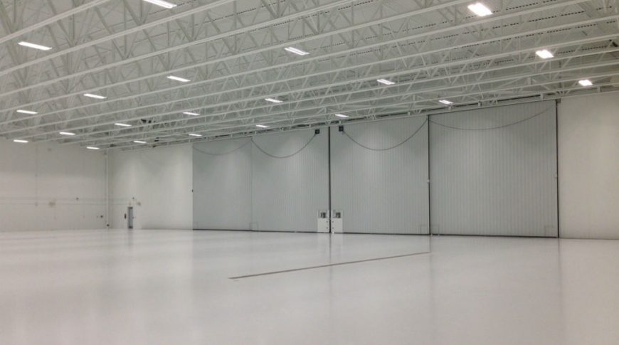 Large empty interior of hangar with white flooring and white metal roof with exposed white trusses and fluorescent lighting overhead with large closed hangar doors to back wall.