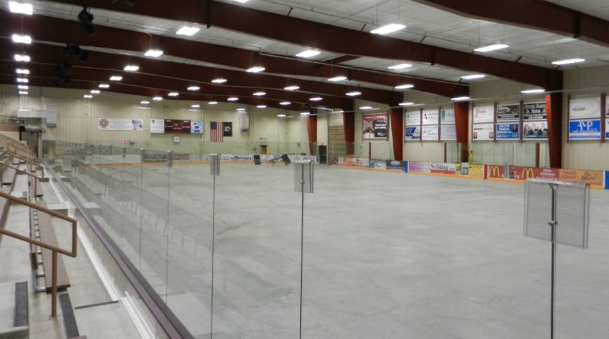 Ice hockey arena with glass and bleacher surround with large brown metal support beams overhead and business banners throughout.