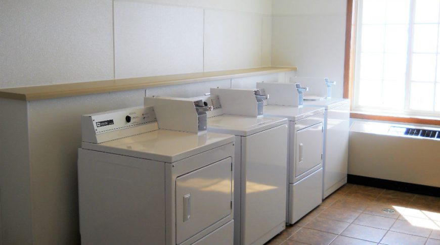 four coin-operated laundry machines in small room with large sunny window in back.