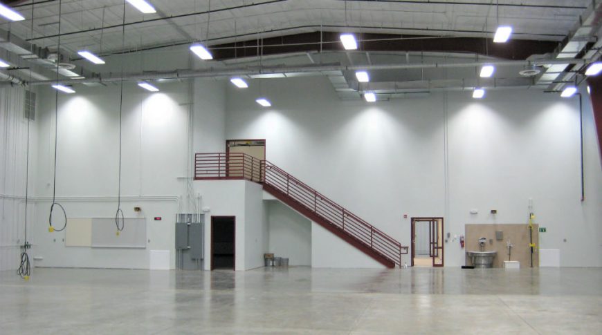 Large, two-story work area with light white walls and concrete flooring with power cords hanging from ceiling and wash station to back wall.