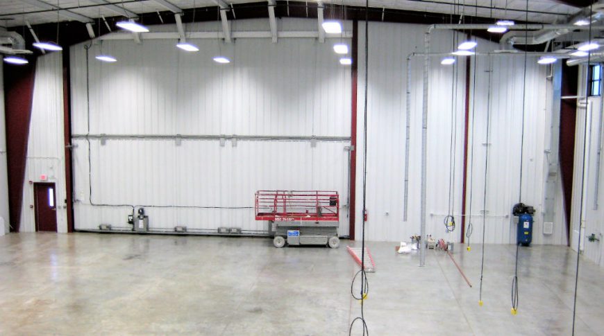 Second-story view of large white metal workshop interior with power cords hanging from ceiling and concrete floors underneath. No workstations installed.