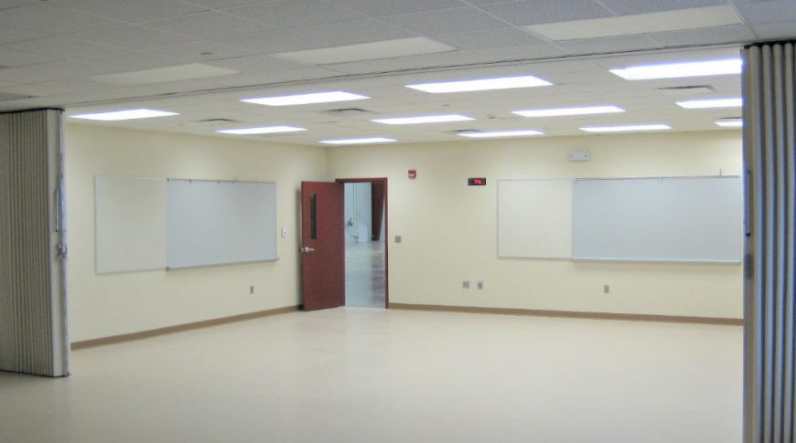 Interior room with beige vinyl flooring, paneled ceiling tiles with fluorescent lights and two whiteboards on back left and right walls with accordion dividers in center of room.