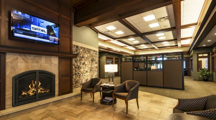 Interior reception area with light beige tiles beneath and hearth with fire to the right surrounded by dark brown wooden millwork and big-screen tv above. Cubicles to the back.