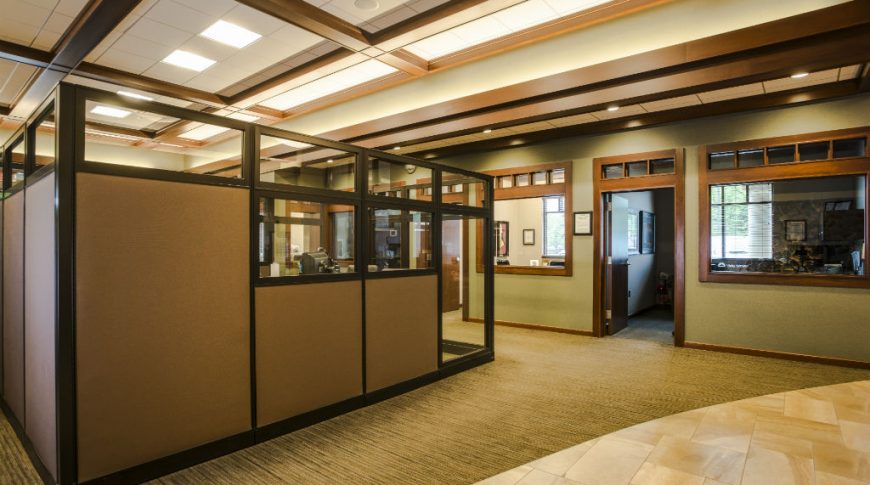 Large room with privacy-paneled cubicles to the center with private offices with doors and windows behind. Dark brown wooden ceiling millwork above.