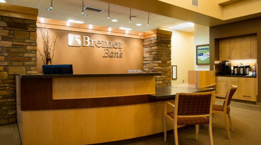 Light-wooden two-level reception desk with dark black and white speckled countertops. Brown wall behind with the metal words "Bremer Bank" and two brick columns flanking each side.