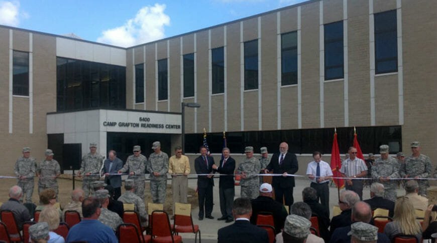 Grand opening ribbon-cutting ceremony after completion of Camp Grafton Readiness Center. Large two-story light brick and window building in the background. Foreground with many people in military uniforms and suits holding ribbon and audience seated in front.