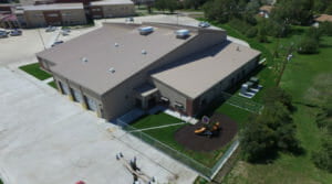 Aerial view of light concrete and red brick building with light metal roof. Outside it is a small fenced in grassy area with a toddler playground at the center.