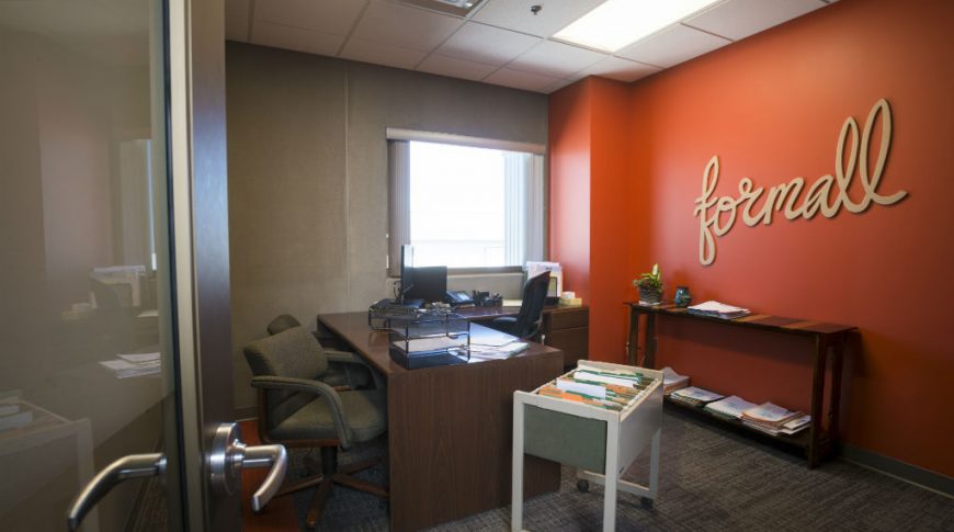 Office room with concrete wall and one window to back left and bright orange back right accent wall with the word "formall" in large cursive letter sign on it. Brown L-shaped desk and a few chair to back wall.