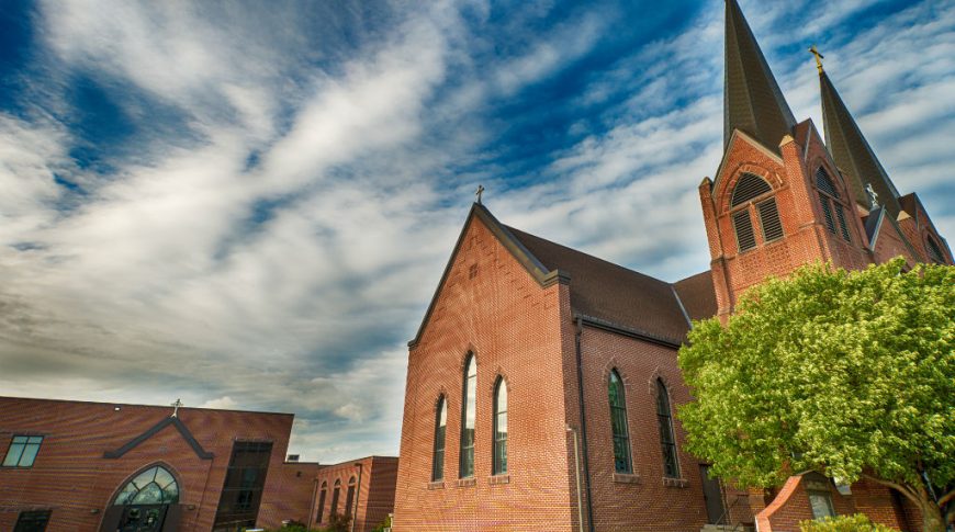Side view of red brick catholic church with grand steeples and stained glass arched windows throughout.