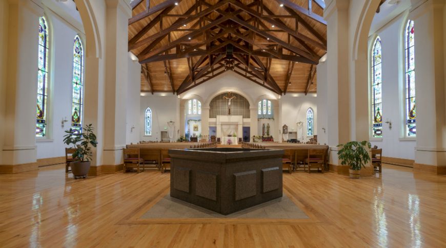 Entrance view of chapel with granite holy water fountain in front surrounded with light wooden floors and wooden pews and pulpit to the background with wooden-paneled ceiling and exposed trusses in the background.