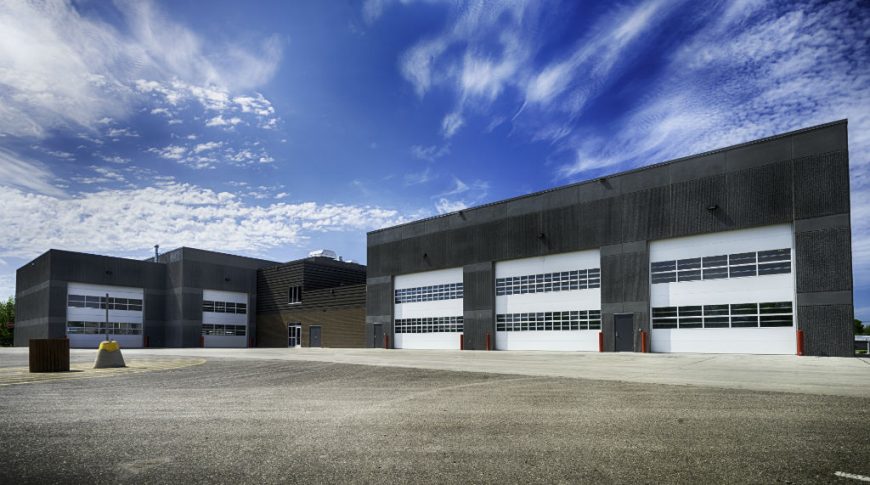 Large equipment garage bays with five large white garage doors with two rows each of windows on dark gray precast concrete building and paved parking lot in foreground.