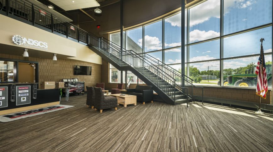 Entryway to NDSCS Bisek Hall building with curved wall of windows to the back, staircase in front leading from second floor mezzanine to ground level reception entryway.