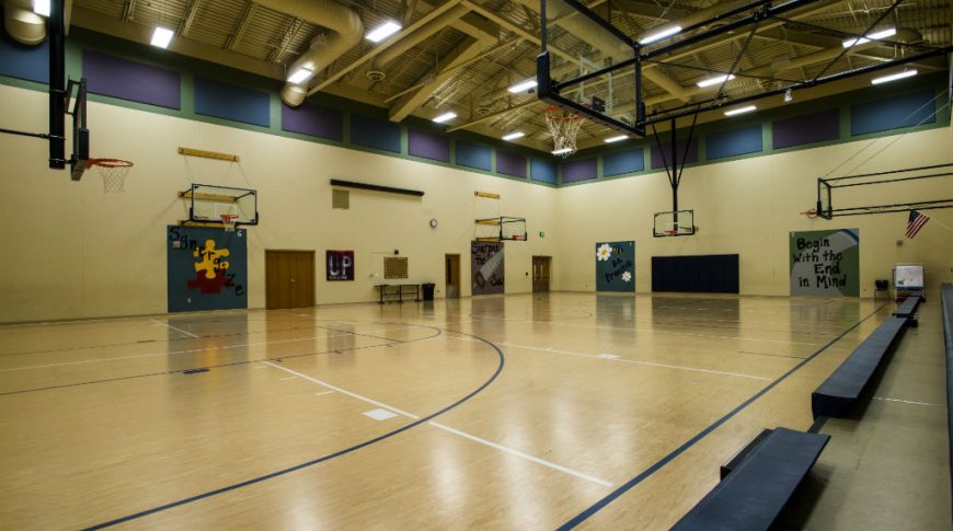 Beige gymnasium with light faux-wood flooring and one wall of low-rise retractable bleachers and some large inspirational posters on each wall.