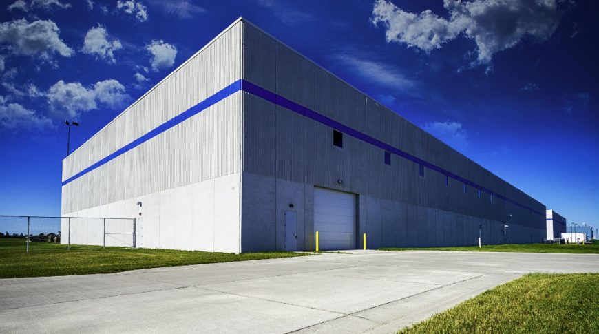 Exterior beige precast building with bright blue stripe along the top third of the building with loading bay and bright blue sky.