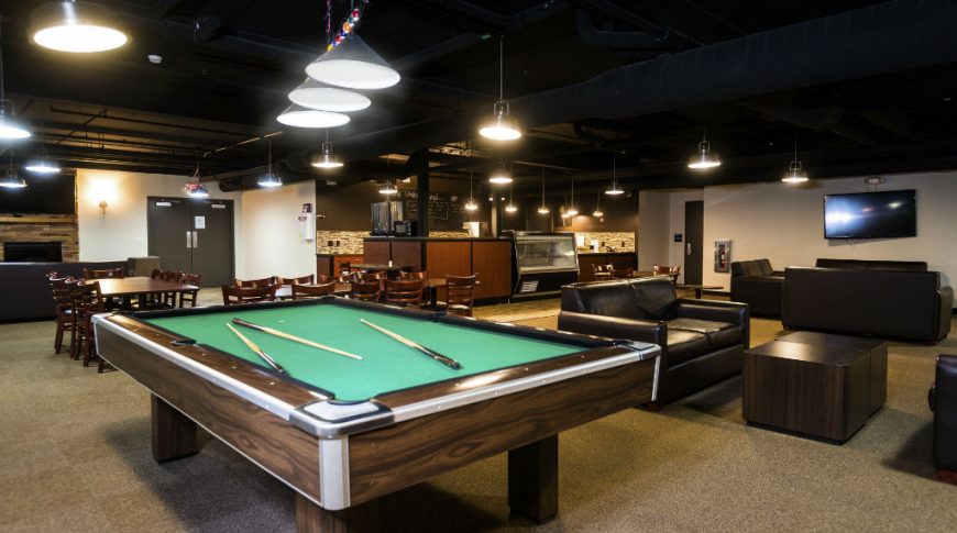 Game/lounge room with large pool table to the foreground with tables and chairs to back left, and black leather coaches and tables to back right with tvs on the back left and right walls. Cafe area to the back center.