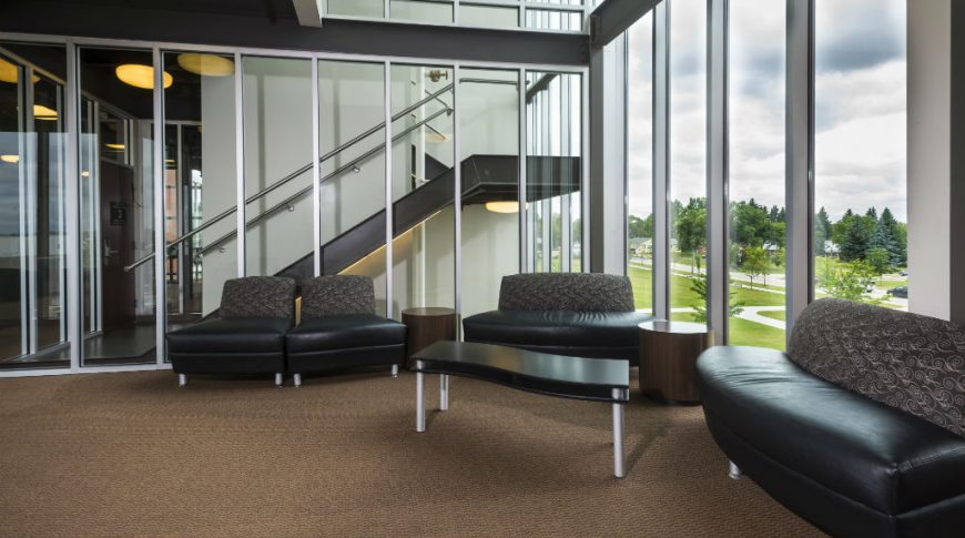 Sitting area with brown carpeted flooring with black curved leather and fabric coaches and accent tables surrounded by walls of floor-to-ceiling windows on left and back side.