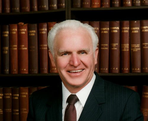 Head and shoulders portrait of owner Lynn Comstock who is an older white male with white hair wearing black suite, white-collared shirt and dark maroon and polka dot tie with library book backdrop