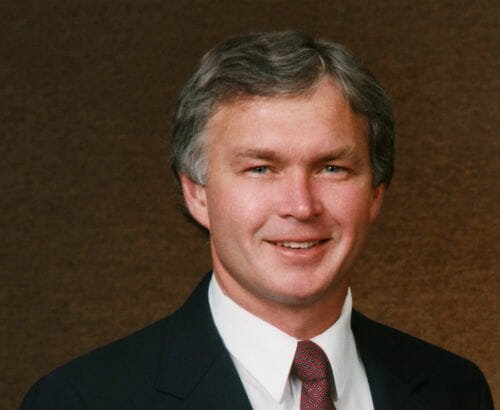 Middle-aged man with salt and pepper hair wearing black suite, white button-up and red speckled tie on brown backdrop