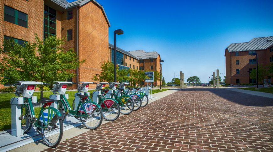 Brown brick bike path with 10 bike rental stations on the left-hand side and brown brick buildings on either side of the path.