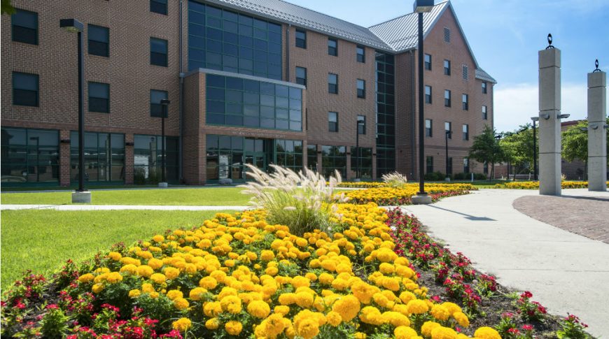Exterior view of four-story brown brick learning-living college center with grassy knoll in front and concrete pathways bordered with bright yellow marigold and other flowers.
