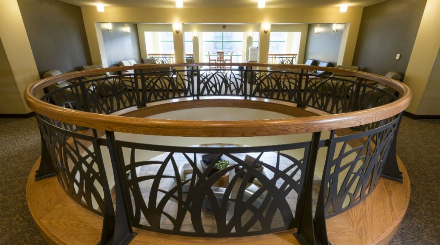 Second floor round rotunda with metal grass-detailed custom railing with wooden handrail opening to seating area below.
