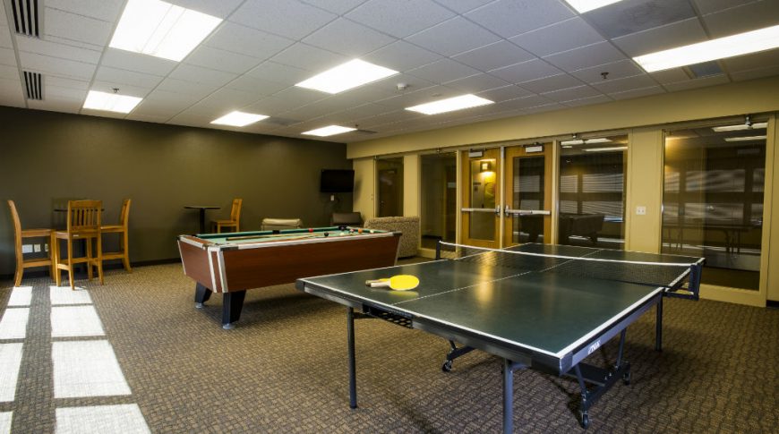 Game room with ping-pong table in foreground and pool table in the middle with some bar-height tables and chair on the back wall.