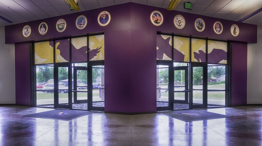 School entryway from the inside looking out the front doors. The back wall is bright purple with medal circles and painted graphics overhead with gold and purple eagles appliques on the tops of the entryway door windows.