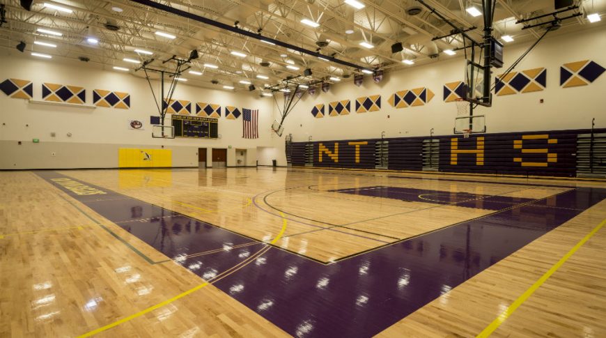 Brightly-lit gymnasium viewing from the corner of the court with basketball hoops lowered on two ends, purple bleachers retracted with the letters "NTHS" in yellow spelled on the bleachers' side. Purple and yellow padded accents on the tops of the walls to buffer noise.