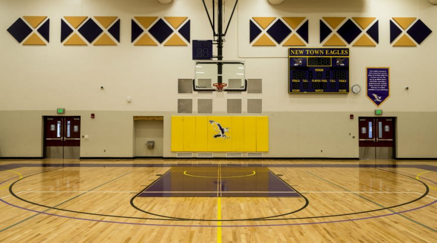 Halfcourt view looking to basketball hoop and backboard wall with purple and gold and eagle accents and scoreboard on the top right of back wall flanked by two double doors on each side.