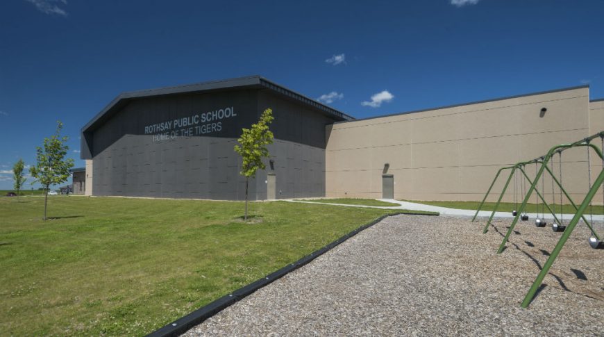 Exterior of brown precast concrete and dark charcoal-colored paneling with the words "Rothsay Public School. Home of the Tigers" on he wall. Playground area with green swings in upper right foreground.
