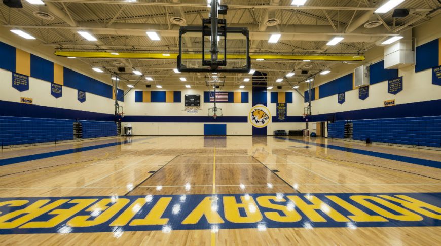 Brightly lit gymnasium with newly polished light oak floors, with rich blue and yellow school colors throughout. Folded bleachers on left and right sides.