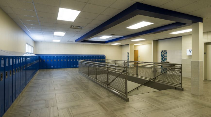 School hallway of blue lockers to back and left walls with stairwell going downstairs in the middle of the room and bathrooms to the back right.