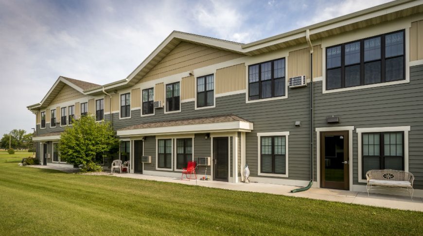 Exterior view of Siena Court Assisted Living building with two-tone grey/green and beige siding with lush green lawn and trees throughout. Showcases exterior doors to private apartments and small concrete patios outside each one.