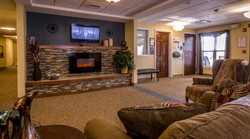 Cozy interior of assisted living with plush fabric coach and armchair with fluffy pillow facing a wide fireplace and tv mounted above.
