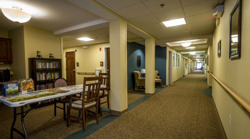 Dark carpeted hallway with beige painted walls and handrails to right side of wall with a dark teal painted sitting nook with two armchairs to back left of the hall and a folding 8-foot table with unfinished puzzle and three chairs around it in a nook on the upper left-hand side of the hall.